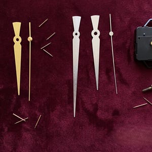 Può includere: Componenti per orologi disposti su una superficie bordeaux. Le parti includono lancette dorate e argentate, piccoli perni, un movimento dell'orologio nero e una vite. Le lancette variano per dimensioni e forma, con una miscela di finiture metalliche.