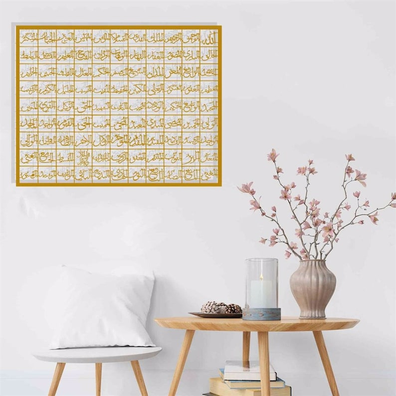 May include: A gold framed wall art with Arabic calligraphy in a white room with a wooden table, a white pillow, and a vase with pink flowers.