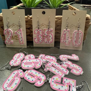 May include: Pink and white number earrings with colorful sprinkles. The numbers are 21, 35, 40, 8, 9, 7, 2, 3, 6, 9, 5, and 7.