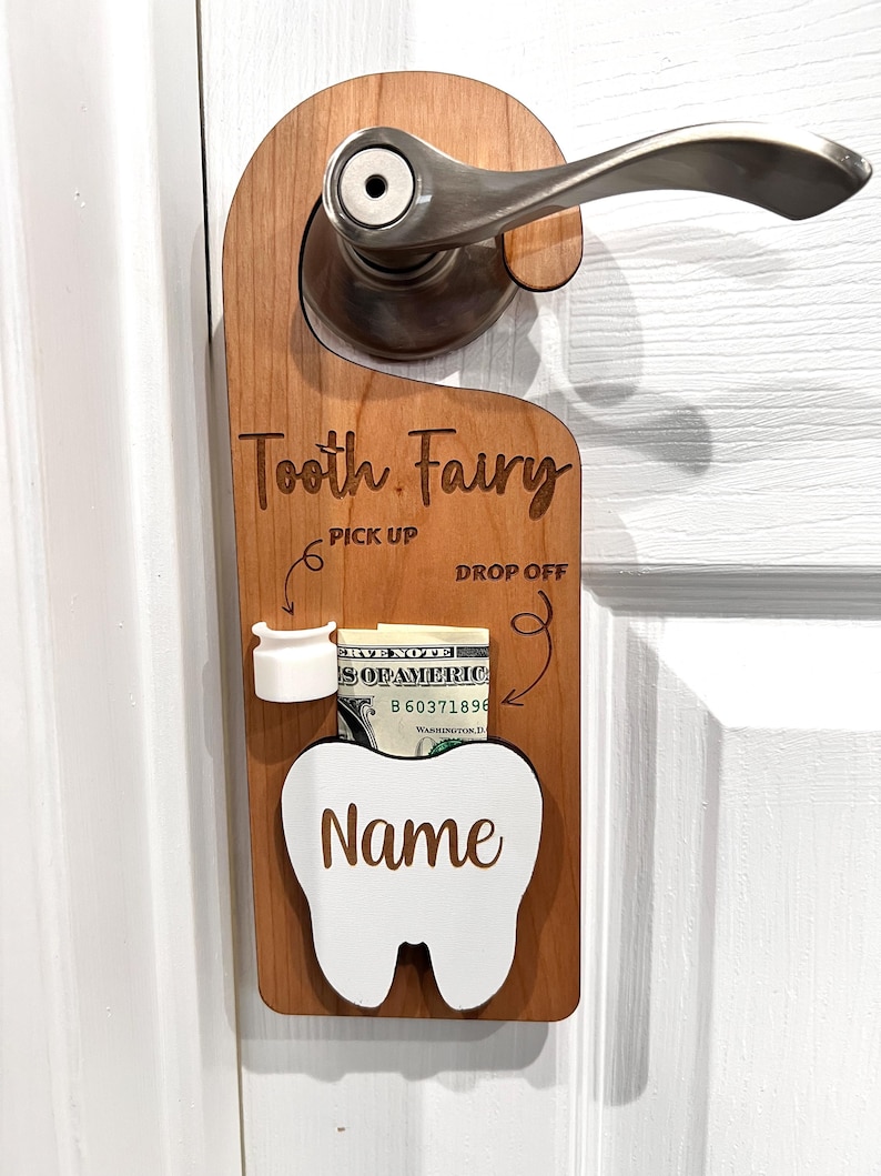 May include: Wooden tooth fairy door hanger with a white tooth cutout, a slot for a tooth, and a space for money. The hanger has the words "Tooth Fairy", "Pick Up", and "Drop Off" written on it.