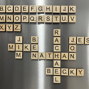 May include: A close-up of a wooden Scrabble game board with the letters "MIKE", "NATHAN", "BECKY", "JESUS", and "RAC" spelled out. The letters are arranged in a crossword puzzle pattern.