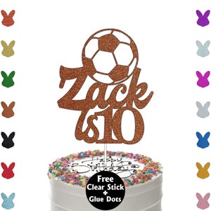 K&ouml;nnte beinhalten: Ein kupferfarbener, glitzernder Tortenaufsatz mit einem Fu&szlig;ball-Design, dem Namen "Zack" und der Zahl "10". Der Aufsatz befindet sich auf einer wei&szlig; glasierten Torte mit bunten Streuseln und dem Schriftzug "Happy Birthday".