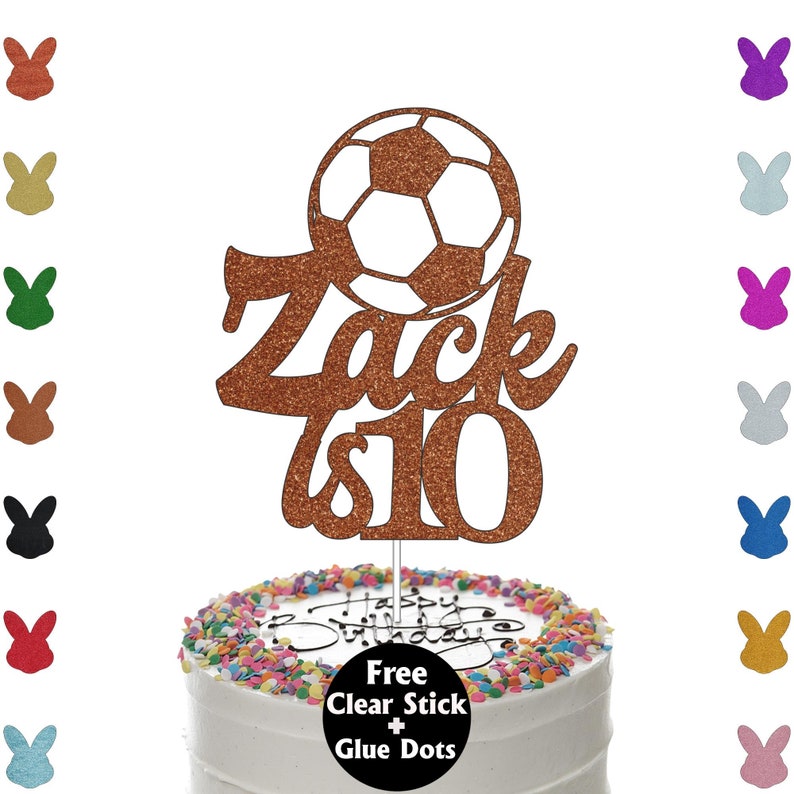 K&ouml;nnte beinhalten: Ein kupferfarbener, glitzernder Tortenaufsatz mit einem Fu&szlig;ball-Design, dem Namen "Zack" und der Zahl "10". Der Aufsatz befindet sich auf einer wei&szlig; glasierten Torte mit bunten Streuseln und dem Schriftzug "Happy Birthday".