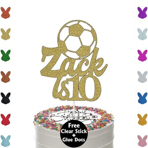 K&ouml;nnte beinhalten: Ein goldener Glitzer-Kuchenaufsatz mit einem Fu&szlig;ball-Design und dem Namen "Zack is 10". Der Aufsatz befindet sich auf einem wei&szlig; glasierten Kuchen mit bunten Streuseln und dem Schriftzug "Happy Birthday".