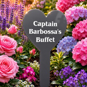 May include: A dark grey heart-shaped garden stake with a pointed bottom, featuring the white text "Captain Barbossa's Buffet" printed in the centre. The stake is positioned upright in dark soil, surrounded by pink roses and purple flowers.