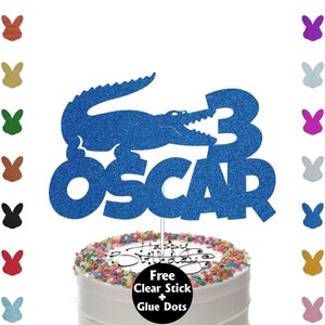 May include: Blue glitter cake topper featuring a crocodile and the name "Oscar" along with the number "3". A free clear stick and glue dots are included.