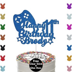 May include: Blue glitter cake topper featuring a video game controller design, the words "Happy 11th Birthday Brody" and two stars. A free clear stick and glue dots are included.