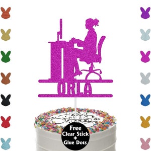 May include: A magenta glitter cake topper depicting a person at a computer, with the name "ORLA" beneath. The topper is on a frosted cake decorated with colourful sprinkles and the words "Happy Birthday".