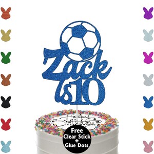 K&ouml;nnte beinhalten: Ein blau glitzernder Tortenaufsatz mit einem Fu&szlig;ball-Design und dem Namen "Zack is 10". Der Aufsatz befindet sich auf einer glasierten Torte mit bunten Streuseln und dem Schriftzug "Happy Birthday".
