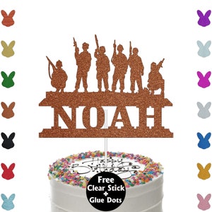 May include: A brown glitter cake topper featuring a silhouette of six soldiers holding guns and the name NOAH. The topper is on a white cake with colourful sprinkles. Free clear stick and glue dots are included.