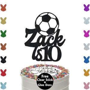 K&ouml;nnte beinhalten: Ein schwarzer Glitzer-Kuchenaufsatz mit einem Fu&szlig;ball-Design, dem Namen "Zack" und der Zahl "10". Der Kuchen ist wei&szlig; mit bunten Streuseln und dem Schriftzug "Happy Birthday".