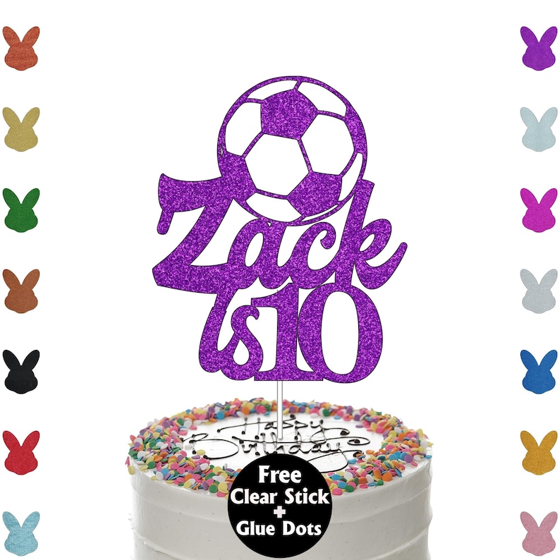K&ouml;nnte beinhalten: Ein lila Glitzer-Kuchenaufsatz mit einem Fu&szlig;ball-Design und dem Text "Zack is 10". Der Aufsatz befindet sich auf einem wei&szlig; glasierten Kuchen, der mit bunten Streuseln verziert ist. Auf dem Kuchen steht "Happy Birthday".