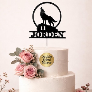 May include: A black acrylic cake topper featuring a silhouette of a wolf howling at the moon, positioned above a rock formation. The design includes the number 11 and the name JORDEN in bold capital letters. A clear stake supports the topper.