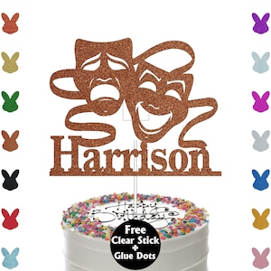 May include: A copper glitter cake topper featuring the comedy and tragedy theatre masks with the name "Harrison". The topper is placed on a white frosted cake with colourful sprinkles and the words "Happy Birthday".