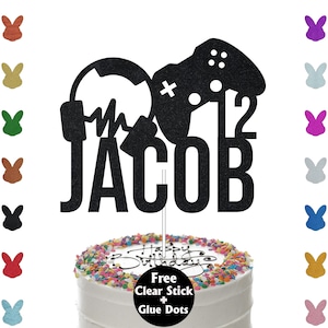 May include: Black glitter cake topper featuring a video game controller, headphones, and the name "Jacob". The topper is displayed on a white cake with colourful sprinkles. Free clear stick and glue dots are included.