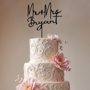 May include: A three-tiered wedding cake with white icing and floral decorations. The cake is decorated with pink flowers and a black cake topper reading "Mr + Mrs Bryant". The cake has an elegant, classic design.