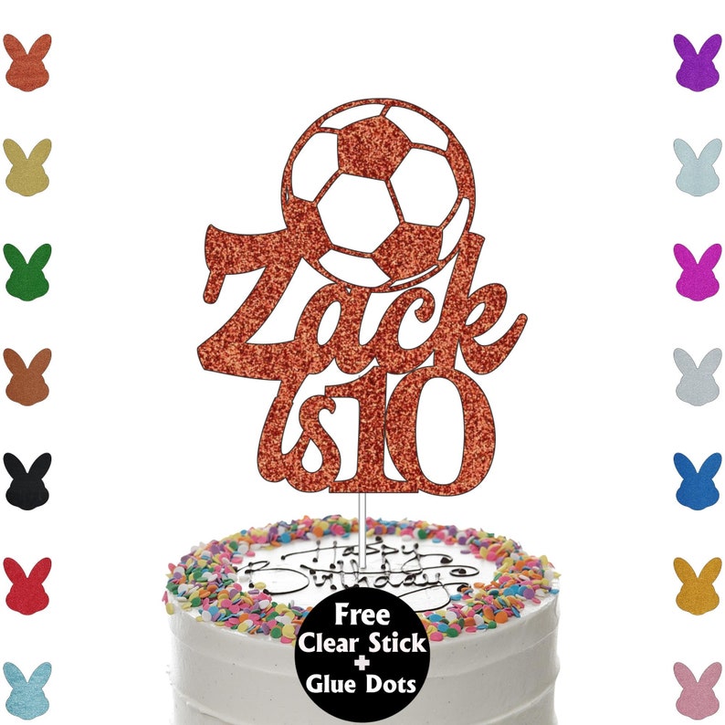 K&ouml;nnte beinhalten: Ein orangefarbener, glitzernder Tortenaufsatz mit einem Fu&szlig;ball-Design und dem Namen "Zack" mit der Zahl "10". Der Aufsatz befindet sich auf einer wei&szlig; glasierten Torte mit bunten Streuseln. Der Text "Happy Birthday" ist sichtbar.
