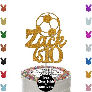 K&ouml;nnte beinhalten: Ein goldener Glitzer-Kuchenaufsatz mit einem Fu&szlig;ball-Design, dem Namen "Zack" und der Zahl "10". Der Kuchenaufsatz befindet sich auf einem wei&szlig; glasierten Kuchen mit bunten Streuseln und den Worten "Happy Birthday".