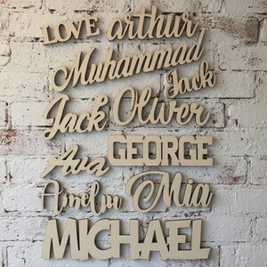 May include: Wooden name cutouts in various fonts, including "Arthur", "Muhammad", "Jack", "Oliver", "George", "Ava", "Amelia", "Mia", and "Michael". The names are displayed against a white brick wall.