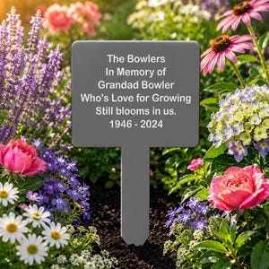 May include: A rectangular grey garden memorial stake with a pointed bottom, featuring white text that reads: The Bowlers, In Memory of Grandad Bowler, Who's Love for Growing, Still blooms in us., 1946 - 2024. The stake is positioned in dark soil.