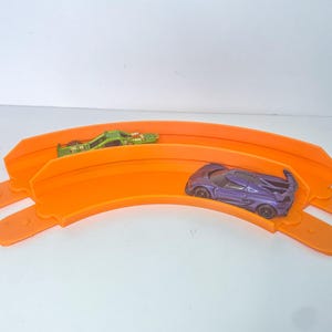 May include: A bright orange toy race track with two toy cars. One car is green, the other is purple. The track is curved and has two lanes. The toy cars are small and appear to be made of metal or plastic.