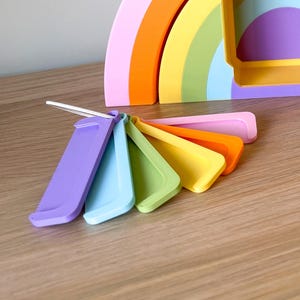 May include: A set of seven rainbow-coloured measuring spoons. The spoons are arranged in a fan shape, with the largest spoon at the top and the smallest spoon at the bottom. The spoons are made of a smooth, plastic material.