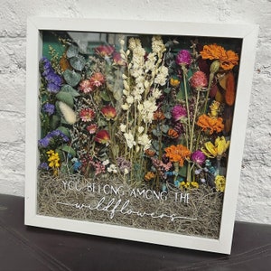 Puede incluir: Una pieza de arte de flores prensadas enmarcada en blanco con una variedad de flores secas y vegetación. El texto "You belong among the wildflowers" está escrito en letras blancas sobre un fondo marrón.