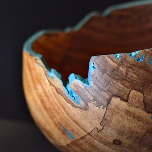 May include: A handcrafted wooden bowl with a unique, irregular edge. The bowl's interior is a warm brown, while the exterior features a natural wood grain. The edge is accented with a bright turquoise blue, adding a pop of color to the piece.