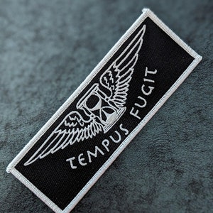Może przedstawiać: Czarna naszywka haftowana z białymi ściegami. Naszywka przedstawia skrzydlaty klepsydrę z łacińskim napisem "Tempus Fugit", co oznacza "Czas ucieka".