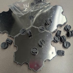 May include: A collection of interlocking, hexagonal metal plates with a shiny, reflective surface. The plates are surrounded by a gray plastic frame. Small, gray plastic connectors and screws are scattered around the plates, with some still in a clear plastic bag.