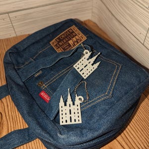 May include: A blue denim bag with a zipper closure and two straps. The bag has a brown leather patch with the text "A Chosen Generation" and a white temple keychain attached.