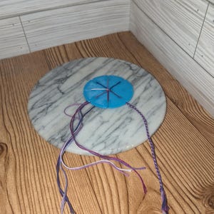 May include: A blue, circular handmade item with purple and white strings attached. The item is on a white and gray marble-like surface, set against a wooden background. The strings are braided and tied at the ends.