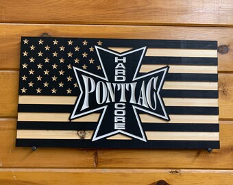 May include: A wooden American flag with a black and white cross design in the center. The cross has the word "Pontiac" in white letters and "Hardcore" in white letters vertically on the cross.