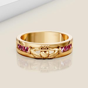 May include: A gold Claddagh ring with pink gemstones. The ring features the traditional Claddagh design of two hands clasping a heart, topped with a crown. The gemstones are set around the band.