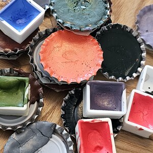 May include: A collection of colorful watercolor paints in small, square white containers. The paints are various shades of blue, green, red, purple, and orange. The paints are set inside of repurposed bottle caps, creating a unique and artistic presentation.