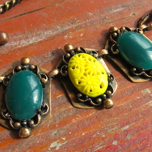 May include: A vintage necklace with a copper-toned chain and pendant settings. The necklace features three oval pendants: two with teal-colored stones and one with a yellow, daisy-shaped design. The necklace is displayed on a rustic, reddish-brown wooden surface.