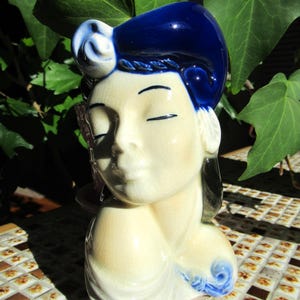 Pode incluir: Estatueta de cerâmica da cabeça e ombros de uma mulher, com os olhos fechados. O cabelo é azul escuro brilhante e a pele é creme. Um detalhe floral azul está no ombro. A estatueta é provavelmente um item decorativo.