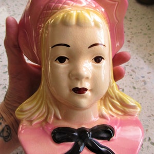 May include: A vintage ceramic planter shaped like a girl's head with a pink bonnet and a black bow. The girl has blonde hair and a painted face.