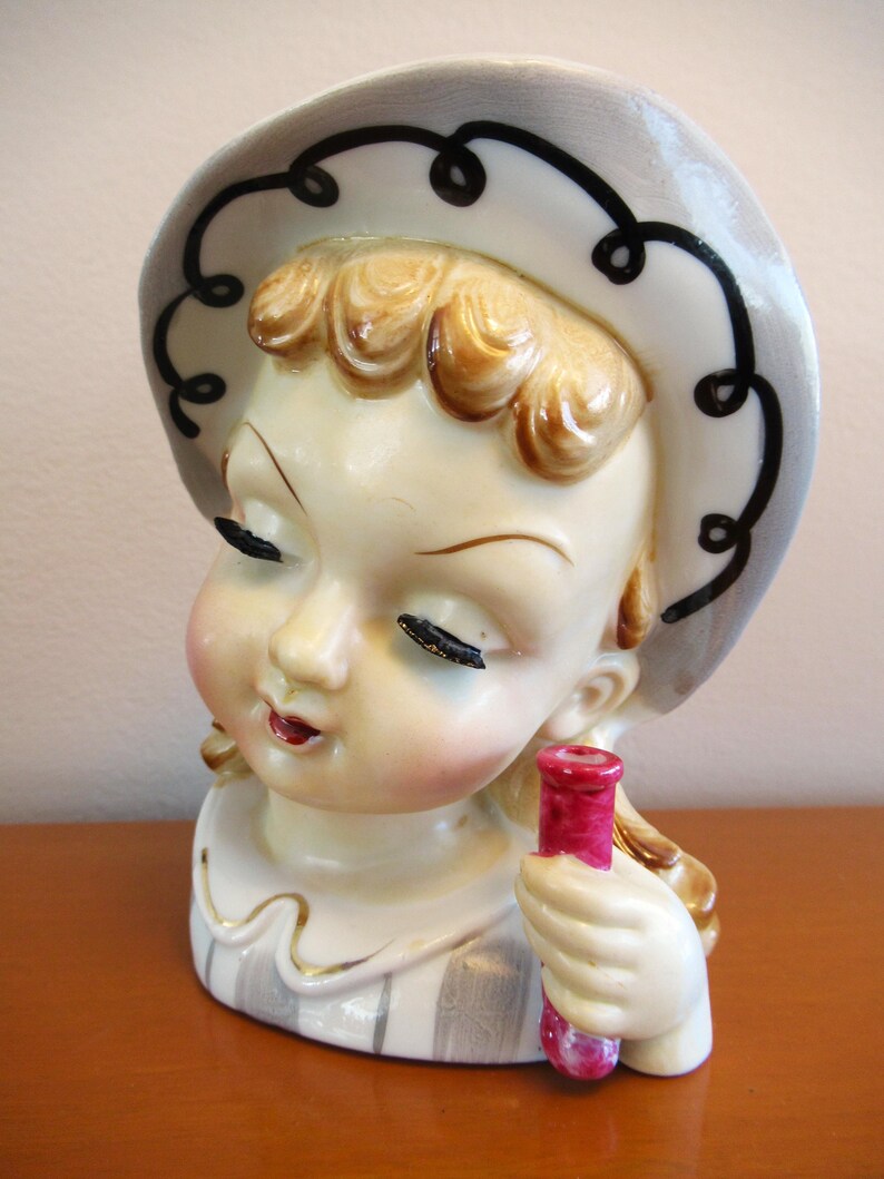 Puede incluir: Jarr&oacute;n de cer&aacute;mica vintage con forma de cabeza de ni&ntilde;a con los ojos cerrados, que lleva un sombrero gris con ribete negro. La ni&ntilde;a tiene el pelo rubio y sostiene un vial rosa. El jarr&oacute;n tiene un dise&ntilde;o a rayas blancas y grises.