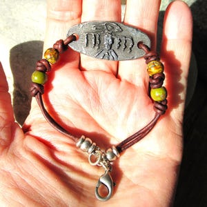 Puede incluir: Una pulsera artesanal con una pieza central de metal plateado con un diseño en relieve. La pulsera incluye cordones de cuero marrón, cuentas decorativas en tonos verdes y amarillos y un cierre plateado. La pulsera mide aproximadamente 17.78 cm de largo.