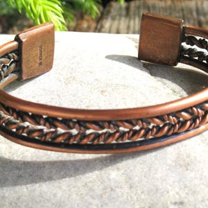 May include: Copper cuff bracelet with a braided design. The bracelet has a polished finish and a hinged closure. The braided pattern is a darker shade of copper, creating contrast. The bracelet is displayed on a light gray stone surface.