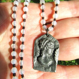 Op de afbeelding: Een ketting met een hanger met een klassieke figuur en een kralenketting. De hanger is rechthoekig met een gedetailleerd reliëf van een gezicht. De ketting heeft witte, ronde kralen geregen op een donker koord.