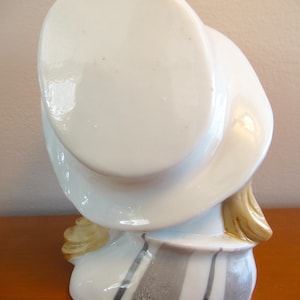 Puede incluir: Figura de cer&aacute;mica de una persona con un sombrero blanco grande, con pelo rubio. La figura tiene un dise&ntilde;o a rayas blancas y grises en la base. El sombrero est&aacute; inclinado, sobre una superficie de madera.