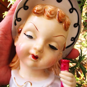 Puede incluir: Un bolsillo de pared de cer&aacute;mica vintage con forma de cara de ni&ntilde;a. La ni&ntilde;a tiene los ojos cerrados, labios rojos y un sombrero blanco con ribete negro. Sostiene un tubo de ensayo rosa. El bolsillo de pared est&aacute; pintado en colores pastel suaves.