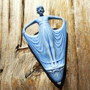 May include: A light blue ceramic wall plaque featuring a stylized figure with arms raised, holding the edges of a flowing gown. The figure has a smooth, glazed finish with visible fine crackle patterns throughout the light blue surface.