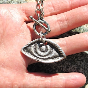 Peut inclure: Un pendentif en métal argenté en forme d'œil avec une finition sombre et texturée. Le pendentif est fixé à une chaîne épaisse et texturée de couleur argentée, dotée d'un fermoir à bascule composé d'un anneau circulaire et d'une barre en T.