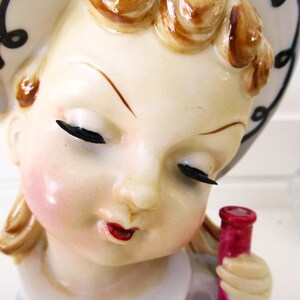 Puede incluir: Figura de cer&aacute;mica vintage de una ni&ntilde;a con los ojos cerrados, mejillas sonrosadas y labios rojos. Lleva un sombrero blanco con espirales negras y sostiene una botella roja. La figura tiene detalles brillantes y una est&eacute;tica cl&aacute;sica.