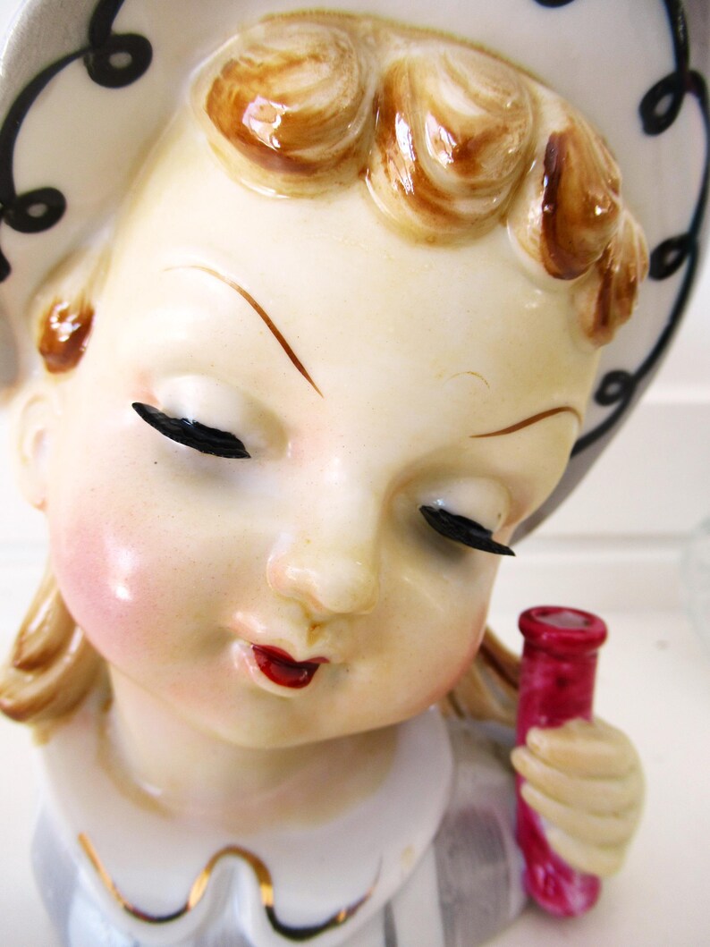 Puede incluir: Figura de cer&aacute;mica vintage de una ni&ntilde;a con los ojos cerrados, mejillas sonrosadas y labios rojos. Lleva un sombrero blanco con espirales negras y sostiene una botella roja. La figura tiene detalles brillantes y una est&eacute;tica cl&aacute;sica.