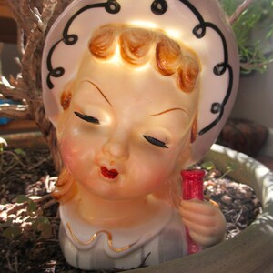 Puede incluir: Figura de cer&aacute;mica vintage de una ni&ntilde;a con los ojos cerrados, con un sombrero con remolinos negros. Sostiene una botella roja. La figura tiene un tono de piel claro, labios rojos y est&aacute; colocada en una maceta.