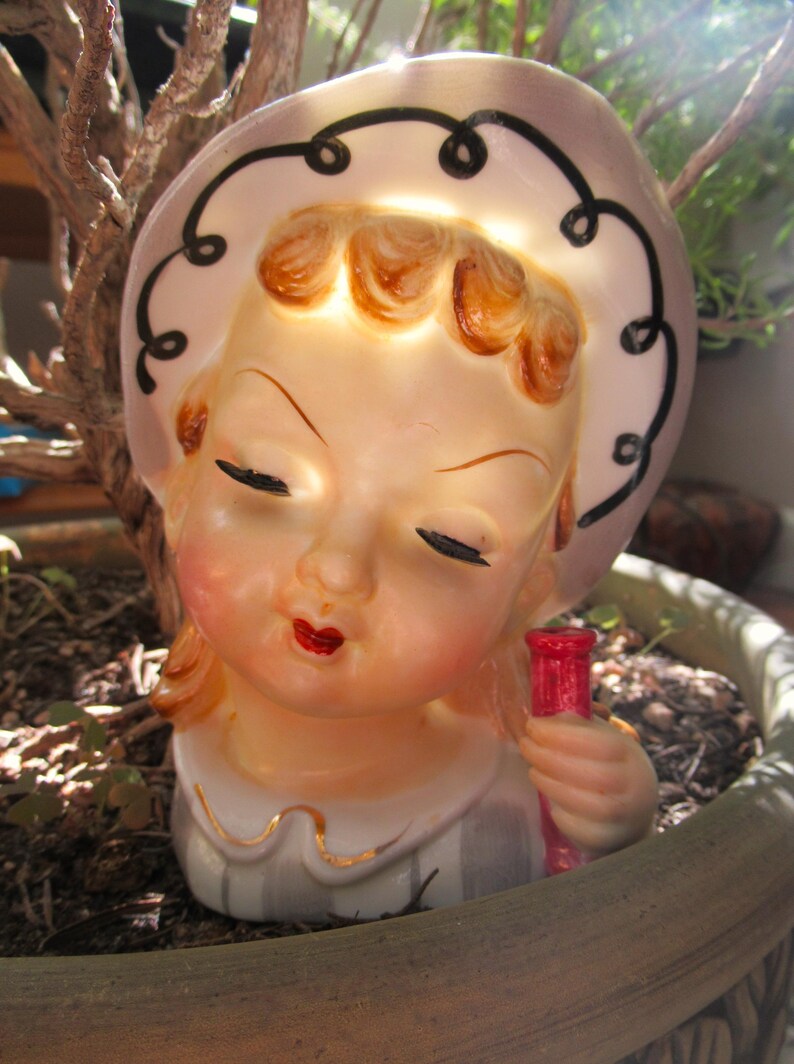 Puede incluir: Figura de cer&aacute;mica vintage de una ni&ntilde;a con los ojos cerrados, con un sombrero con remolinos negros. Sostiene una botella roja. La figura tiene un tono de piel claro, labios rojos y est&aacute; colocada en una maceta.